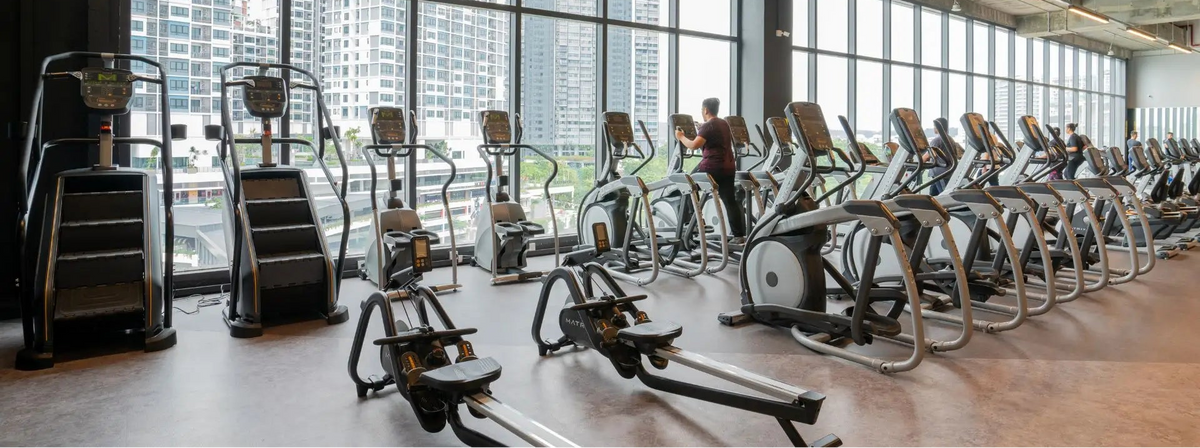 A row of cardio exercise machines, including treadmills and stair climbers, arranged in a modern gym with large windows overlooking a city skyline.
